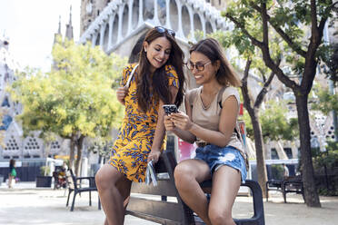 Frauen, die ein Mobiltelefon benutzen, während sie auf einer Bank vor der Sagrada Familia in Barcelona, Katalonien, Spanien sitzen - JSRF01174