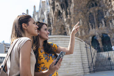 Lächelnde Frau, die auf einen Freund zeigt, der ein Mobiltelefon an der Sagrada Familia in Barcelona, Katalonien, Spanien, benutzt - JSRF01188