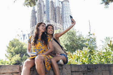 Lächelnde Freunde machen ein Selfie, während sie auf einer Stützmauer im Sagrada Familia Park sitzen, Barcelona, Katalonien, Spanien - JSRF01193