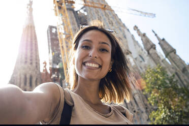 Lächelnde Frau, die ein Selfie macht, während sie vor der Sagrada Familia in Barcelona, Katalonien, Spanien steht - JSRF01194