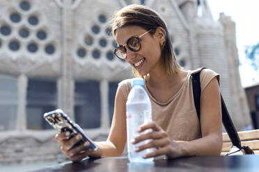 Junge Frau mit Sonnenbrille, die ein Mobiltelefon benutzt, während sie vor der Sagrada Familia in Barcelona, Katalonien, Spanien sitzt - JSRF01195