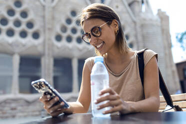 Young woman wearing sunglasses using mobile phone while sitting against Sagrada Familia at Barcelona, Catalonia, Spain - JSRF01195