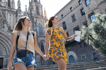 Freundinnen halten sich an den Händen, während sie gegen den Platz der Kathedrale von Barcelona in Barcelona, Katalonien, Spanien, laufen - JSRF01204