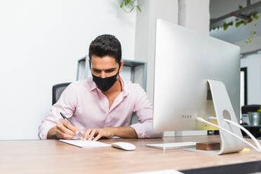 Anonymous concentrated businessman in mask taking note on paper near desktop computer while working on project - ADSF17866