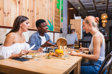 Positive junge multirassische Freunde essen leckere Gerichte und Burger in einem modernen Restaurant - ADSF17872