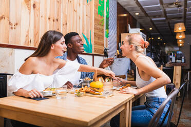 Positive young multiracial friends eating tasty dishes and burgers while gathering in modern restaurant - ADSF17873