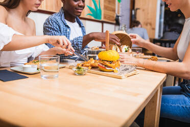 Positive young multiracial friends eating tasty dishes and burgers while gathering in modern restaurant - ADSF17874