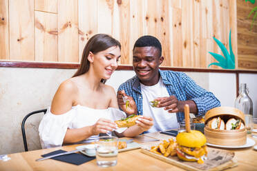 Positive junge multirassische Freunde essen leckere Gerichte und Burger in einem modernen Restaurant - ADSF17876