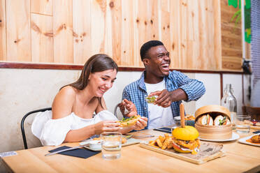 Positive young multiracial friends eating tasty dishes and burgers while gathering in modern restaurant - ADSF17877