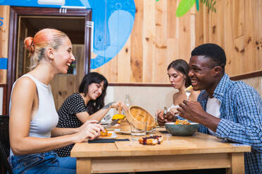 Positive young multiracial friends eating tasty dishes and burgers while gathering in modern restaurant - ADSF17878