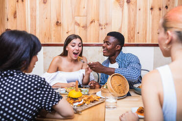 Excited multiracial friends wearing stylish outfits gathering at table in restaurant and spoon feeding each other with tasty fresh dishes - ADSF17879