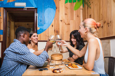 Eine Gruppe fröhlicher multiethnischer Freunde stößt mit ihren Weingläsern an, während sie in einer kreativen Cafeteria ein leckeres Abendessen zu sich nehmen - ADSF17883