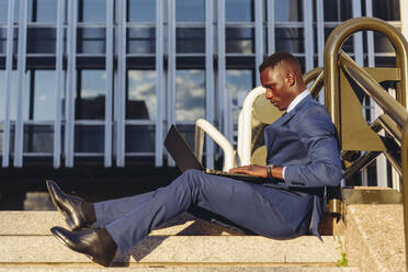 Ganzkörper-Seitenansicht eines konzentrierten afroamerikanischen Finanzmanagers in einem eleganten blauen formellen Anzug, der auf einer Treppe in der Nähe eines modernen Gebäudes sitzt und Nachrichten auf einem Laptop liest - ADSF17905