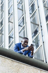 Niedriger Winkel von selbstbewussten respektablen afroamerikanischen Geschäftsmann in formalen Outfit lehnt auf der Straße gegen moderne Gebäude in der modernen Innenstadt mit Smartphone - ADSF17911