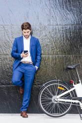 Serious male executive in formal suit leaning on wall while surfing internet on cellphone and standing with hand in pocket near electric bicycle - ADSF17917