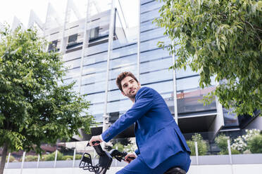 Young smiling man in elegant suit riding electric bicycle on pavement while looking away - ADSF17919
