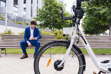 Gutaussehender Geschäftsmann in eleganter Kleidung auf einer Holzbank sitzend, während er die Zeit in der Nähe eines elektrischen Fahrrads in der Stadt überprüft - ADSF17920
