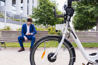 Handsome businessman in elegant clothes sitting on wooden bench while checking time near electric bike in town - ADSF17920