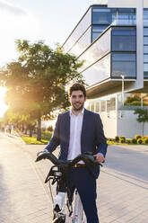 Trendy unshaven male office worker in formal wear strolling on street with electric bicycle while looking at camera - ADSF17921