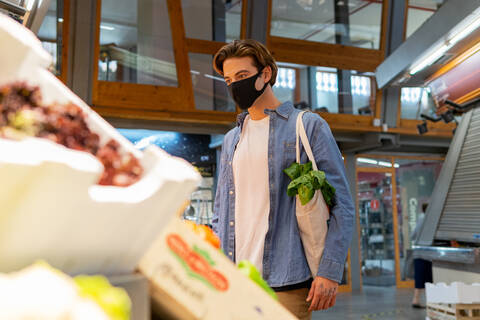 From below young male customer in protective mask carrying eco friendly recycled fabric shopping bag and choosing fresh groceries while buying food in supermarket stock photo