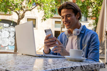 Positiver junger männlicher Fernarbeiter in Freizeitkleidung mit Kopfhörern im Nacken, der eine Nachricht auf seinem Smartphone liest, während er mit Laptop und einer Tasse Kaffee am Tisch sitzt und an einem Projekt auf der Terrasse eines Cafés arbeitet - ADSF17926