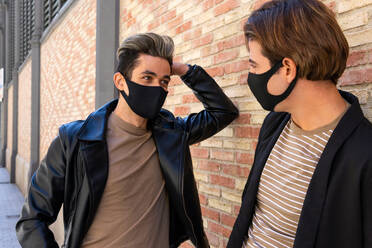 Crop anonymous homosexual friends in stylish wear and face masks looking at each other near wall on street during COVID 19 pandemic - ADSF17931