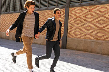 Crop excited gays in trendy apparel holding hands while running on tiled pavement in town near wall and looking forward in back lit - ADSF17935