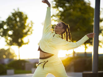 Trendy junge ethnische Tänzerin mit stilvollen Frisur in activewear Durchführung Freestyle-Bewegungen im Park bei Sonnenuntergang - ADSF17942