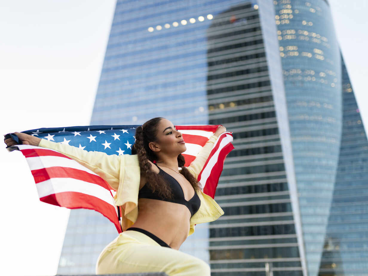 Side view of confident young ethnic lady in sports bra waving American flag  and looking away against modern glass skyscraper stock photo