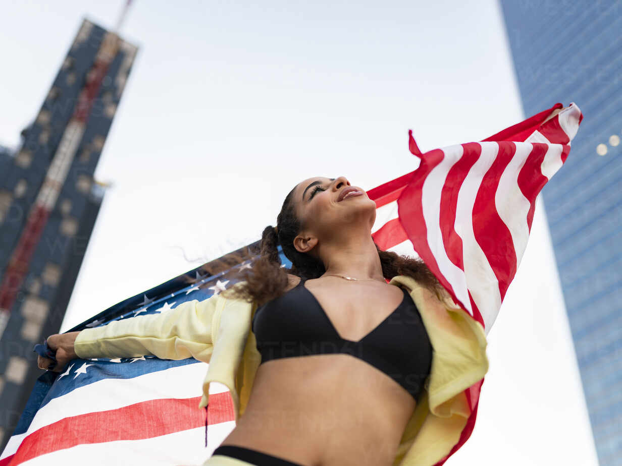 Side view of confident young ethnic lady in sports bra waving American flag  and looking up against modern glass skyscraper stock photo