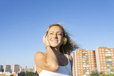 Optimistische junge Frau mit lockigem Haar in weißem Sport-BH, die Musik über Kopfhörer hört, während sie an einem sonnigen Sommertag vor einem unscharfen Stadthintergrund steht - ADSF17968