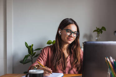 Glückliche junge Frau mit kabellosen Ohrhörern und Brille sitzt am Tisch mit einer Tasse Heißgetränk und tippt auf einem modernen Netbook zu Hause - ADSF17972
