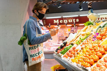 Young male customer in protective mask carrying eco friendly recycled fabric shopping bag and choosing fresh groceries while taking picture on smartphone buying food in supermarket - ADSF17974