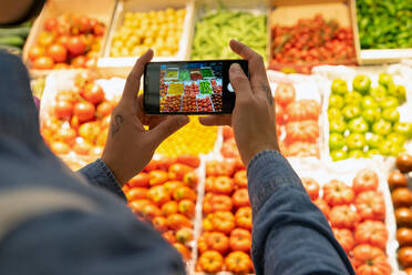 Rückansicht eines anonymen männlichen Käufers, der in der Nähe eines Verkaufsstandes mit Lebensmitteln steht und frisches Gemüse mit dem Handy fotografiert - ADSF17975