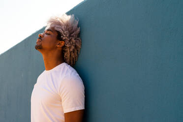 Schwarzer Mann mit Afro-Haar in weiß gekleidet lehnt gegen eine blaue Wand. Afro-Haar-Konzept. Schwarzer Mann in weißem T-Shirt. - ADSF17990