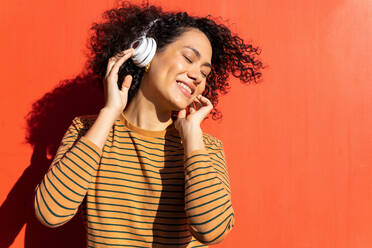 Happy trendy ethnic female with closed eyes and Afro hairstyle enjoying song in headset on red background - ADSF17995