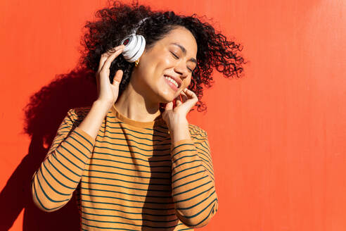 Happy trendy ethnischen Frau mit geschlossenen Augen und Afro-Frisur genießen Song in Kopfhörer auf rotem Hintergrund - ADSF17995