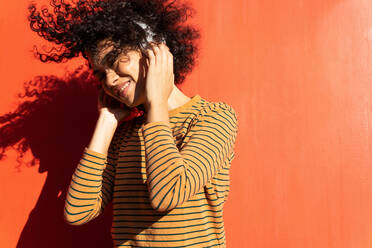 Happy trendy ethnischen Frau mit geschlossenen Augen und Afro-Frisur genießen Song in Kopfhörer auf rotem Hintergrund - ADSF17996