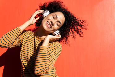 Happy trendy ethnic female with closed eyes and Afro hairstyle enjoying song in headset on red background - ADSF17997