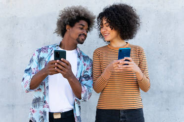 Content multiracial couple of partners in trendy clothes with Afro hairstyle standing near wall with cellphones - ADSF18009