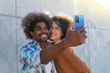 Inhalt multirassischen Paar von Freunden mit Afro-Frisur, die selfie auf Handy, während die Umarmung in der Nähe von Wand im Sonnenlicht in den Rücken beleuchtet - ADSF18010