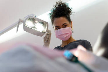 From below of cheerful black female doctor adjusting dental lamp in modern bright clinic before treatment - ADSF18022