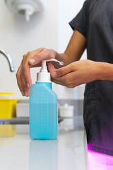 Faceless female doctor in uniform disinfecting hands with antibacterial gel in plastic bottle placed on table in medical room - ADSF18024