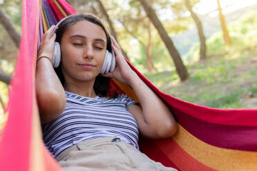 Peaceful female in headphones lying in hammock and enjoying calm music with closed eyes - ADSF18030