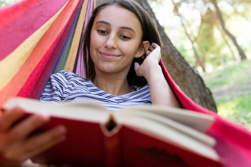 Tranquil female lying in hanging hammock in park and enjoying interesting book at summer weekend - ADSF18033