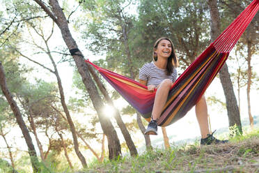 Low angle of young glad female sitting in hammock hanging between trees in garden while enjoying summer and looking away - ADSF18035