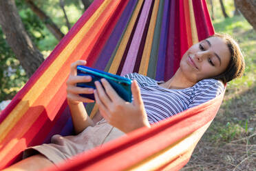 Ausruhende Frau, die in der Hängematte liegt und einen Film auf dem Handy anschaut, während sie ein Wochenende im Sommer im Park verbringt - ADSF18036