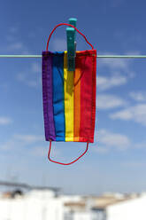 Schutzmaske mit Bild der LGBT-Regenbogenflagge, die mit einer Wäscheklammer an einem sonnigen Tag im Hinterhof befestigt wird - ADSF18039