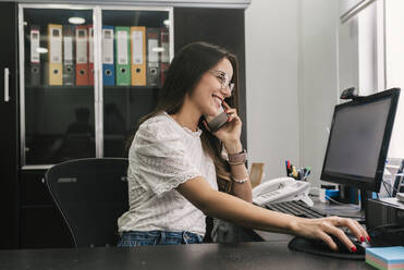 Smiling businesswoman working on computer while talking on mobile phone at office - DSIF00212