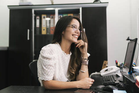 Thoughtful businesswoman smiling while sitting with hand on chin at office stock photo